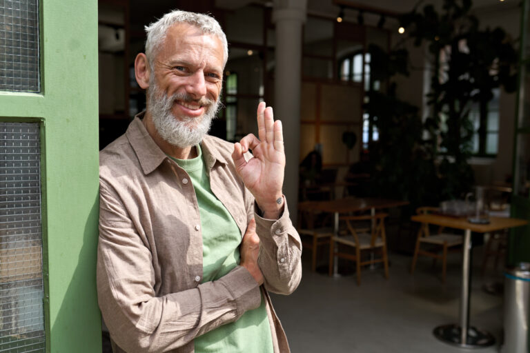 Happy older man small business owner showing ok sign standing outside cafe