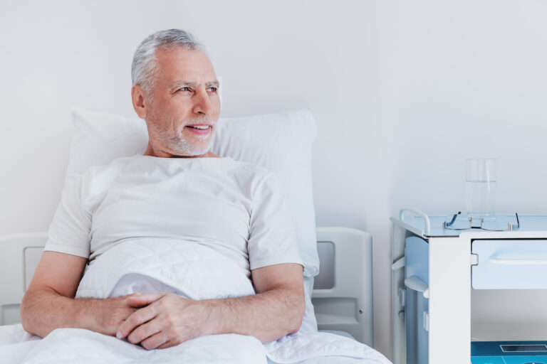 Male senior patient relaxing in bed in hospital ward while looking in window