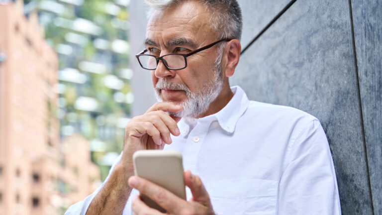 Old senior professional business man holding phone using smartphone thinking